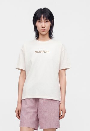 Mujer con cabello negro, vestida con una camiseta blanca de Napapijri y pantalones cortos texturizados de color lila claro, de pie contra un fondo blanco liso.