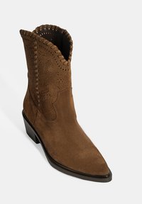Braune Wildleder-Ankle-Boots mit spitzer Zehenform, durchbrochenen Details, gesteppten Akzenten entlang des Kragens und einem geschichteten Absatz.