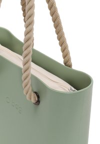 Bolsa O verde con bordes redondeados, exterior texturizado y asas de cuerda trenzada beige. Presenta un bolsillo interior de lona con cremallera.