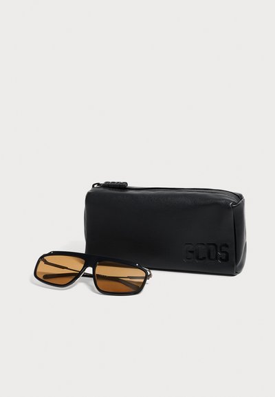 Lunettes de soleil rectangulaires noires avec verres marron à côté d'une pochette noire zippée embossée du logo GCDS sur un fond clair.