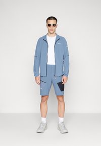 Lys blå treningsjakke med svarte detaljer, kombinert med matchende shorts med en mørk flekk. Blitt brukt med en hvit skjorte og grå joggesko.