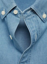 Camicia in denim blu chiaro con colletto a punta, bottoni bianchi e dettagli di cucitura visibili. Il tessuto appare morbido con una texture liscia.