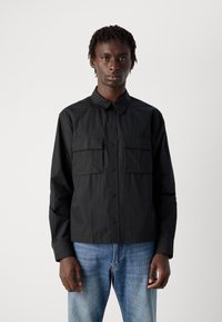 Svart knapp-up skjorte laget av lett materiale, med to brystlommer, krage og lange ermer, sammen med blå jeans.