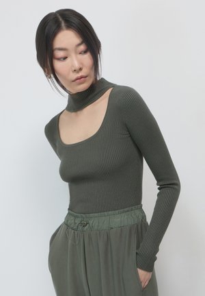 Femme portant un haut en olive vert à manches longues et à col découpé, avec un pantalon à cordon de serrage assorti, regardant vers sa gauche.