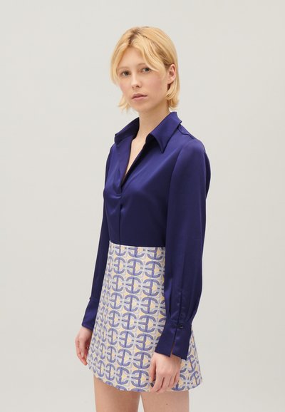Blusa di raso blu navy con maniche lunghe e colletto a punta, abbinata a una minigonna a fantasia in tonalità chiare con accenti blu.
