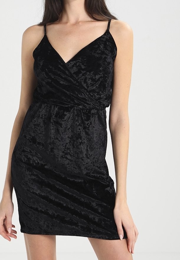 Mini vestido de terciopelo negro con escote en V, finos tirantes tipo espagueti y un diseño envuelto. La tela tiene una superficie suave y texturizada.