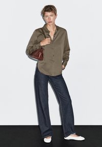 Blouse olive verte à col boutonné avec des manches longues, associée à un jean en denim foncé à jambes larges et des ballerines blanches. Sac marron porté sur l'épaule.