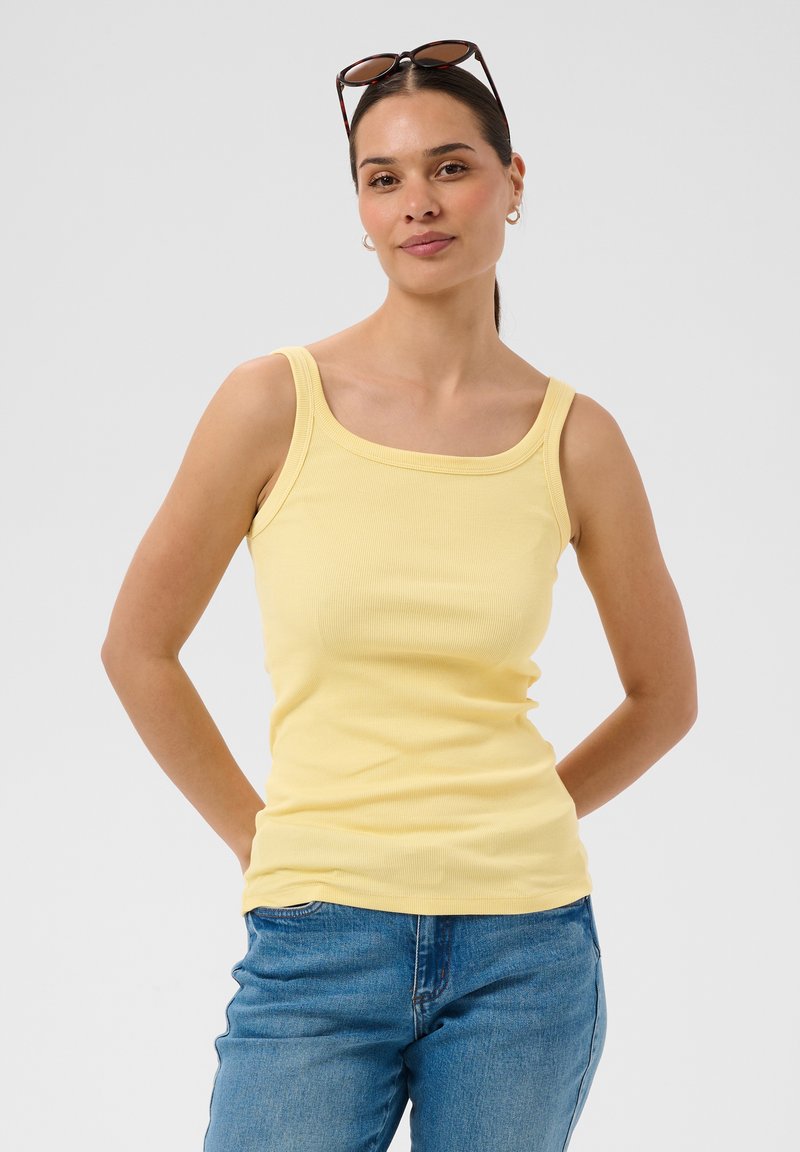 Femme aux cheveux foncés attachés, portant des créoles, des lunettes de soleil sur la tête, un haut jaune sans manches et un jean bleu, debout les mains derrière.