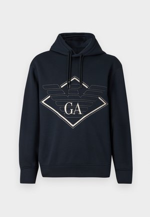Fekete kapucnis pulóver, amelynek elején egy nagy, kontúrozott gyémánt és stilizált szárnylogó látható, rajta az "GA" betűkkel.