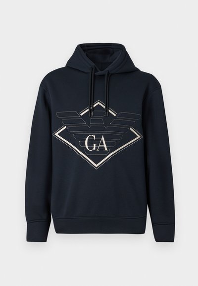 Črna kapuca s kapuco z velikim obrisanim diamantom in stiliziranim logotipom krila, ki vsebuje črki "GA" na sprednji strani.