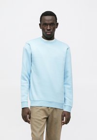 Homme aux cheveux courts et bouclés portant un sweat-shirt bleu clair et un pantalon beige, debout devant un fond blanc uni.