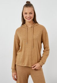Tan-huppari, joka on valmistettu pehmeästä ja teksturoidusta kankaasta, jossa on pitkät hihat ja säädettävät nyörit. Yhdistetty matching-housujen kanssa.