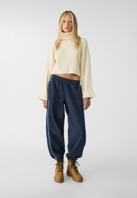 Pull court en tricot côtelé crème, pantalon taille haute bleu marine avec des rayures blanches sur les côtés, bottines à lacets marron. Texture de tissu lisse et coupe décontractée.