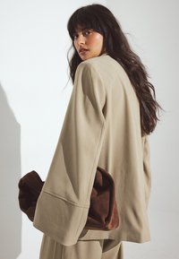 Beige oversized blazer med lange ærmer, glat tekstur; brun ruskinds taske gemt under armen, blød og rund form, minimalistisk design.