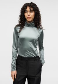 Εφαρμοστή μπλούζα με ζιβάγκο από μεταλλικό πράσινο ύφασμα, με μακριά μανίκια και ομαλή, γυαλιστερή υφή, συνδυασμένη με μαύρο, καλοραμμένο παντελόνι.