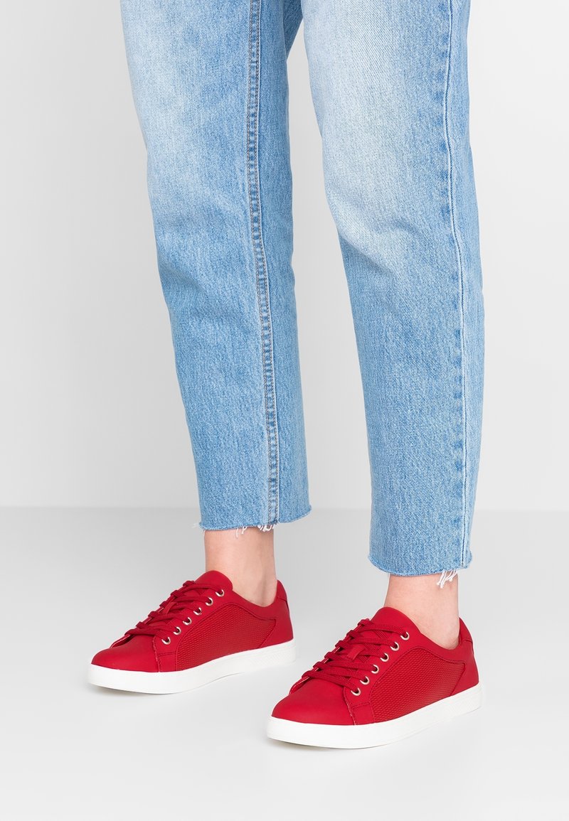 Baskets rouges avec une tige en maille et des accents lisses, avec des semelles en caoutchouc blanches et des œillets ronds pour les lacets, associées à un jean court bleu clair.