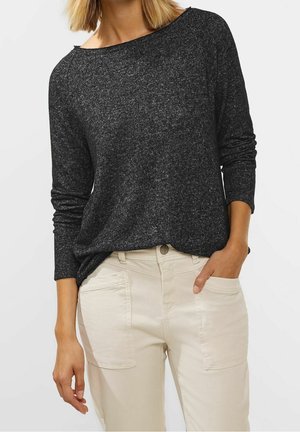 Femme portant un pull à manches longues gris foncé et un pantalon beige clair avec des poches plaquées à l'avant, main dans la poche, torse visible.
