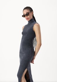 Αμάνικο γκρι φόρεμα με ψηλό λαιμό και πλάγιο σκίσιμο, φτιαγμένο από μαλακό ύφασμα. Συνδυάζεται με γωνιώδεις, σκούρες ηλιακά γυαλιά και πλεξούδα.