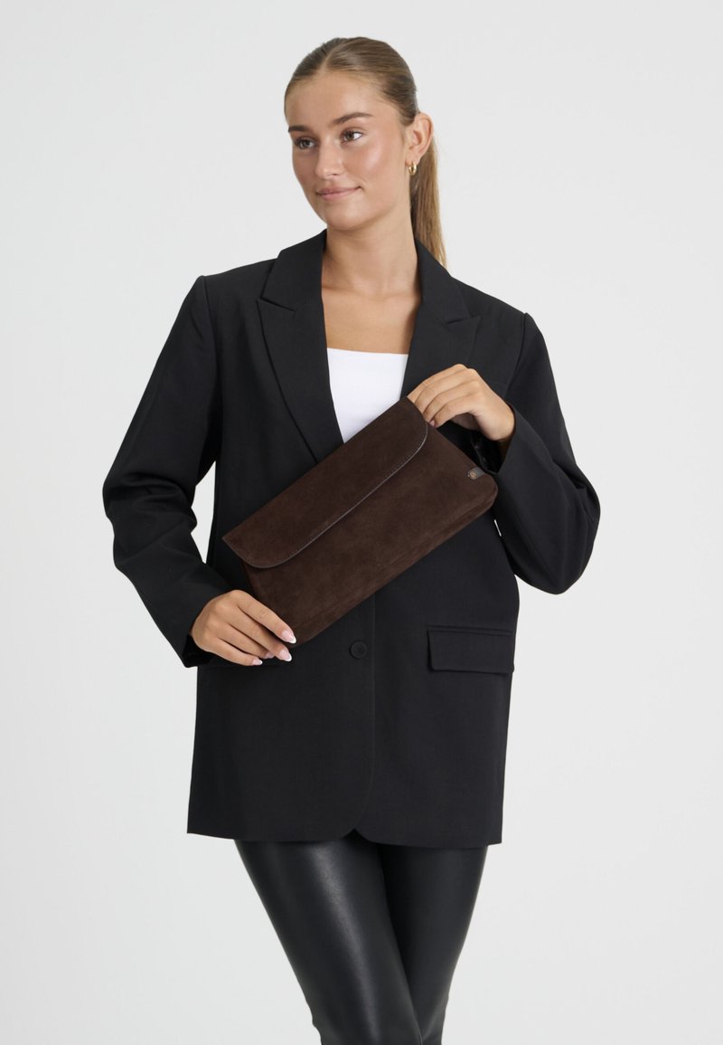 Femme portant un blazer noir et un pantalon en cuir tient une pochette en daim marron sur fond blanc.