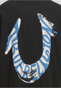 Czarny t-shirt z dużym, abstrakcyjnym, U-kształtnym nadrukiem w kolorach niebieskim i białym, o nakrapianej fakturze i wyraźnym napisie.