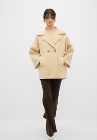Cappotto beige doppiopetto con maniche larghe, dotato di colletto a rever e tasche frontali, abbinato a pantaloni marroni. Tessuto liscio.