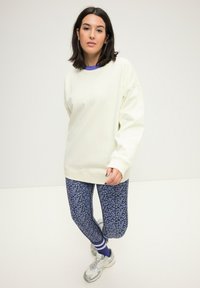 Sudadera oversized de color beige claro con puños de canalé, combinada con leggins azules con un patrón floral. Zapatillas blancas con detalles plateados.