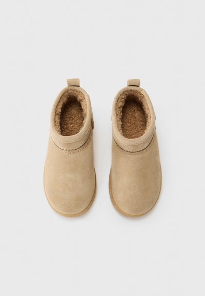 Beige ankelstøvler i suede med en blød, brun pelsforing og en lille trækklap bagpå. Flade såler med minimal detaljer.