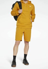 Sweat à capuche jaune moutarde et short assorti en tissu doux. Le sweat à capuche est doté d'une poche kangourou et d'un détail logo. Sac à dos et chaussures noirs.
