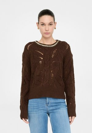 Donna con un maglione a treccia marrone scuro con accento metallico sul collo e jeans azzurri su uno sfondo bianco semplice.