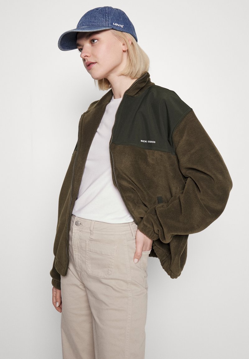 Giacca bomber in pile verde oliva con una spalla in nylon nero, dotata di zip frontale, tasche laterali e logo. Abbinata a una camicia chiara e pantaloni beige.