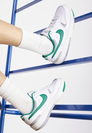 Witte sneakers met een groene Nike-swoosh en afwerking, gedragen met witte geribbelde kousen die rusten op blauwe metalen stangen.