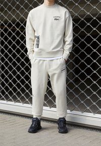 Personne portant un sweat-shirt Lacoste couleur crème et un pantalon de jogging assorti avec des baskets noires, debout devant un fond en grille métallique.