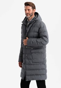Cappotto imbottito grigio con cappuccio, chiusura con zip e bottoni a pressione. Presenta una tasca laterale e una texture trapuntata. Design lungo e dritto.
