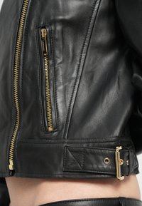 Svart skinnjacka med guldfärgade blixtlås, med en sidoficka med dragkedja, justerbar spänne i fållen och slät textur.