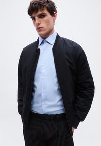 Νέος άνδρας που φοράει ανοιχτό μπλε πουκάμισο με κουμπιά, μαύρο μπουφάν μπόμπερ και μαύρο παντελόνι με ρίγες, στέκεται μπροστά σε μια απλή λευκή φόντο.