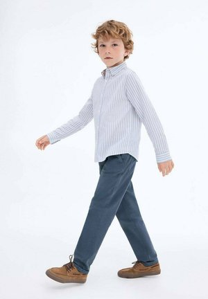 Ragazzino con i capelli ricci rossi che cammina, indossando una camicia bianca a righe con bottoni, pantaloni blu e scarpe marroni su uno sfondo bianco.