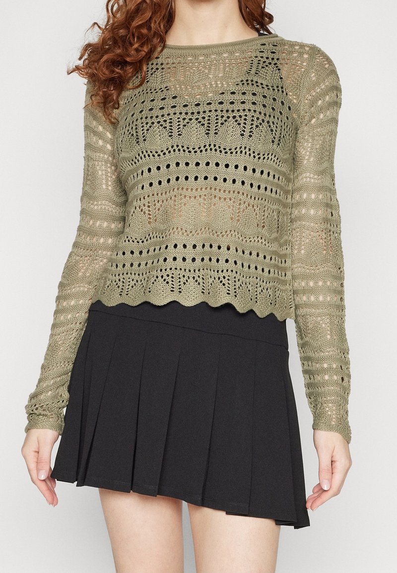 Woman wearing a light olive green crochet knit long sleeve top over a black pleated mini skirt, standing against a plain background.