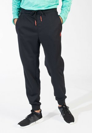 Underkrop iført sorte joggerbukser med orange snøretipper og sorte sneakers med hvide såler, stående med hænderne i lommerne.