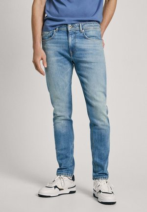 Jeans de mezclilla azul claro con un ajuste slim, diseño de cinco bolsillos y un ligero desgaste. Combinados con zapatillas blancas y negras. Con frente plano.