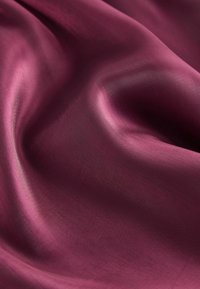 Bordeaux-seidener Stoff mit glatter, glänzender Textur, die in sanften Wellen fällt. Das Licht spiegelt sich von seiner Oberfläche und verstärkt die reiche Farbe.