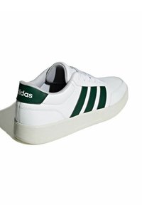 Zapatilla deportiva blanca con detalles en verde, que presenta un diseño de tres franjas, parte superior de cuero liso, puntera redondeada, cierre de cordones y suela acolchada.