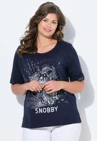 Nainen, jolla on laineikkaat ruskeat hiukset, pukeutuneena tummansiniseen paitaan, jossa on valkoinen koira-illustratio ja "SNOBBY"-teksti, yhdistettynä valkoisiin housuihin.