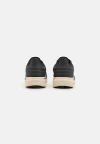 Baskets de sport noires avec une tige lisse, semelle intermédiaire beige rembourrée et semelle extérieure noire texturée. Présente un logo sur le talon.