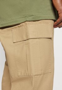 Khaki cargobroeken hebben een gestructureerde stof met een flap op de achterzak. Bovenop zit een olijfgroen shirt dat aan de naad blendt met een gladde afwerking.