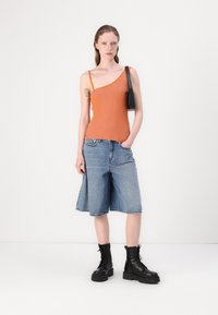 Oranje geribd tanktop met een asymmetrisch ontwerp, gecombineerd met blauwe denim shorts en zwarte veterlaarzen. Inclusief zwarte schoudertas.
