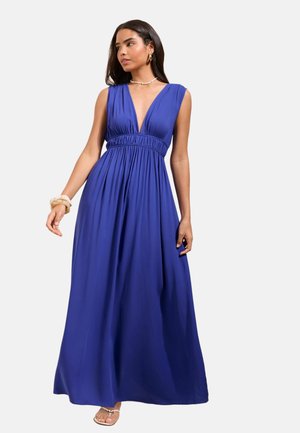 Femme portant une robe longue bleue sans manches avec un décolleté profond en V et une taille haute, accessoirisée avec des boucles d’oreilles, un collier, un bracelet et des sandales.