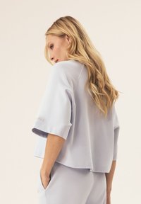 Kobieta z długimi blond włosami nosi luźną, jasnopurpurową bluzkę i dopasowane spodnie, pozując z jedną ręką w kieszeni na jednolitym tle.