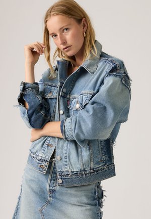 Mujer rubia con chaqueta vaquera de gran tamaño bicolor y falda vaquera a juego, posando con un brazo cruzado y la mano cerca del rostro.