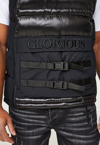 Nahaufnahme einer schwarzen gepolsterten Weste mit dem Schriftzug "GLORIOUS" und zwei horizontalen Schnallenriemen, die über einem weißen Hemd und dunklen Jeans getragen wird.