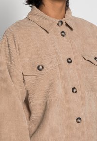 Hellbeige Cordjacke aus Kord mit geripptem Muster, zwei Fronttaschen und rundem Kragen. Mit dunklen Knöpfen und legerer Passform.